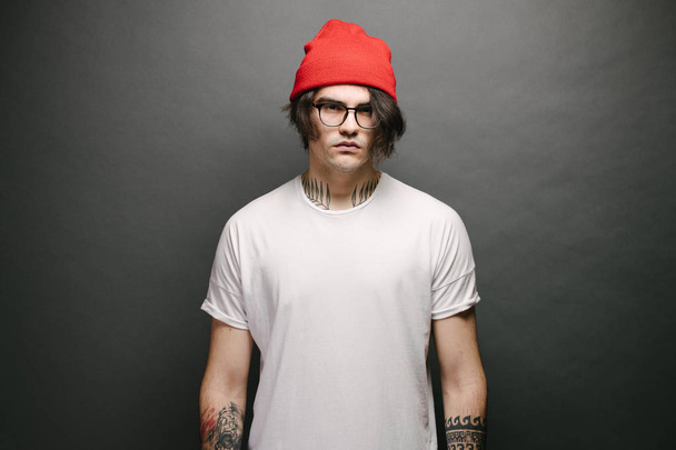 Hipster jóképű férfi modell szemüveg visel fehér üres póló és piros nadrág és egy piros kalap helyet a logó vagy design szürke háttér - Fotó, kép