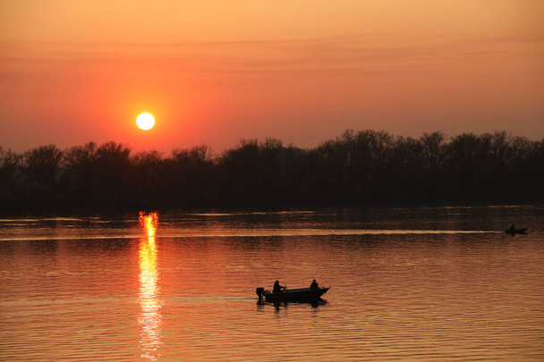 Twee vissers, een man en een vrouw, zwemmen in een motorboot op de rivier tegen de achtergrond van een oranje zonsondergang. Romantische sfeer voor liefhebbers. Goedenavond vissen. Reizen over water. Een natuurlijk fenomeen - Foto, afbeelding