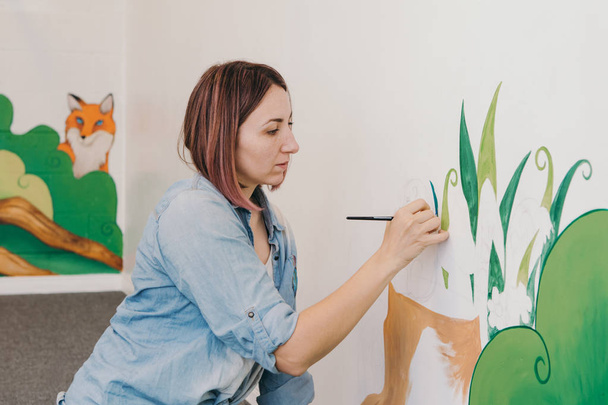 Lifestyle creative hobby and freelance artistic work side job concept. Caucasian woman artist hand painting murals on walls indoor at apartment or studio school with acrylic paints.  - Photo, Image