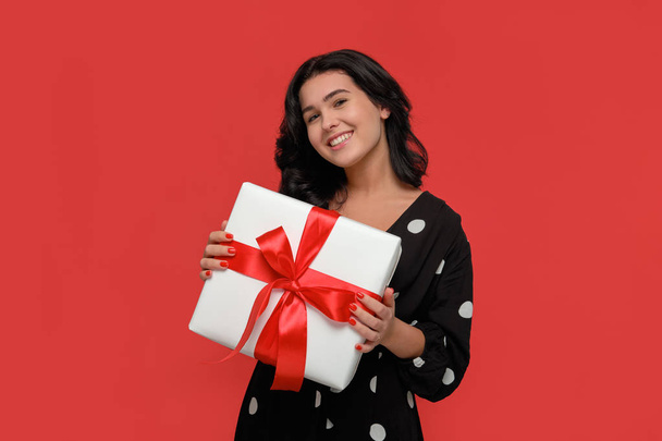Brunette woman in a black dress smiling holding Christmas giftbox of white color with red ribbon. - Fotografie, Obrázek