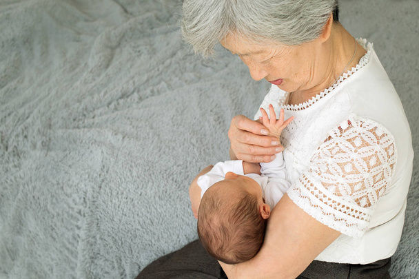 Grandmother admires her sweet granddaughter. Great-grandmother plays with a newborn great-granddaughter. The baby lies in the arms of the grandmother - Photo, Image