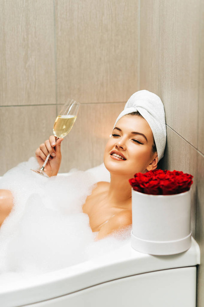 Happy beautiful woman relaxing in a bubble bath tub, holding glass of wine, white towel on the head - Valokuva, kuva