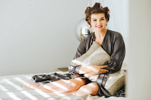 Retrato interior de la joven feliz relajándose en el dormitorio, vistiendo camisón gris, rizadores en la cabeza, sosteniendo una almohada
 - Foto, Imagen