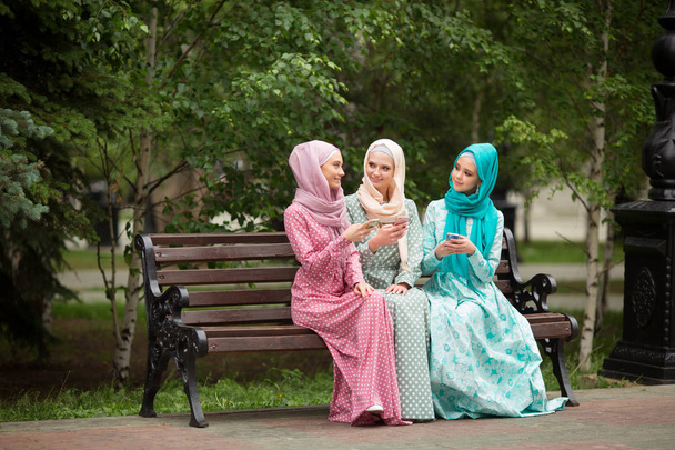 three beautiful young women in muslim dresses in the summer in the park with phones in their hands - Foto, imagen