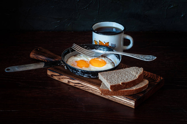 Fried scrambled eggs in a vintage pan, with bread, metal mug with tea - Photo, Image