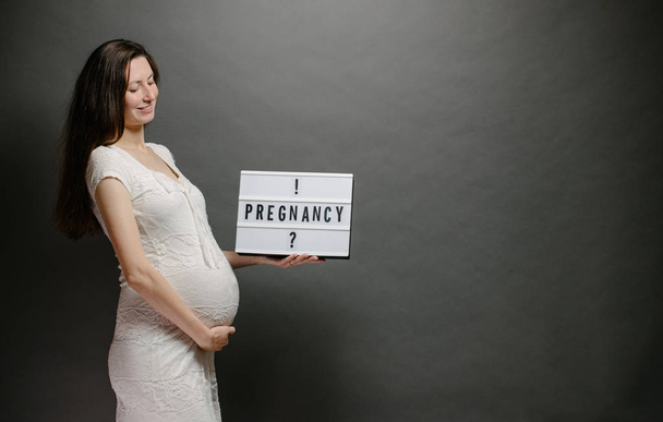 Pregnant woman holding a lightbox with a sign pregnant or pregnancy over gray wall - Foto, Bild