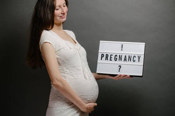 Pregnant woman holding a lightbox with a sign pregnant or pregnancy over gray wall - Photo, Image