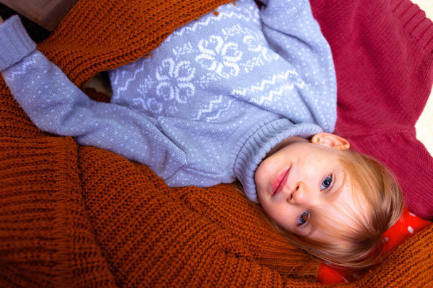 Portrait of a cute little girl in a sweater smiling and lying . Lies in the background of many cozy sweaters. - Foto, Bild