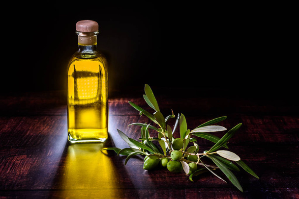 Bottle with virgin olive oil next to a bouquet of raw olives on  - Фото, изображение