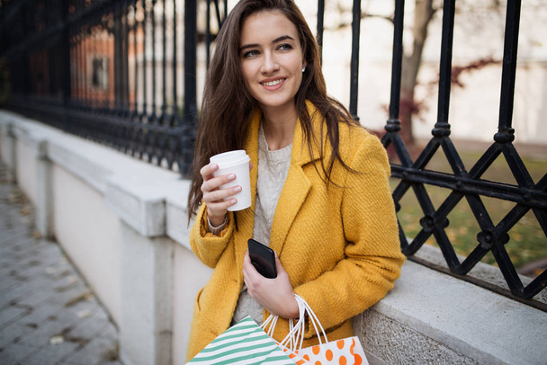 Женщина в желтом пальто пьет утренний кофе и пользуется мобильным телефоном
 - Фото, изображение