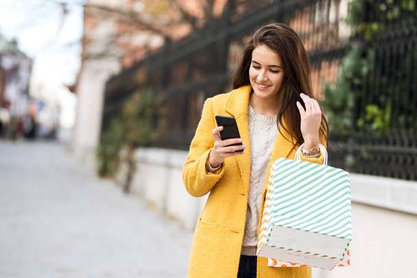Uśmiechnięta kobieta w żółtym płaszczu korzystająca z telefonu komórkowego i torby na zakupy - Zdjęcie, obraz