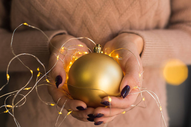 Una chica con un suéter rosa sostiene y da una bola de Navidad dorada en sus manos con una guirnalda amarilla y luces. Hermoso teñido. El concepto de Navidad, Año Nuevo, calidez, celebración
 - Foto, imagen