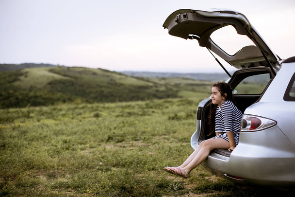 Dívka sedí v otevřeném kufru auta a dívá se na přírodu - Fotografie, Obrázek