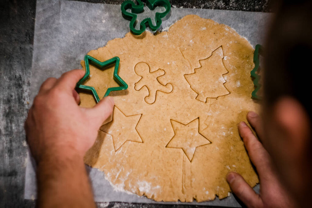 le mani maschili ritagliano il biscotto di pan di zenzero nella forma di un fiocco di neve, un albero di Natale, un uomo da pasta cruda su carta da forno di pergamena su uno sfondo scuro. Vista dall'alto. risparmiare spazio
 - Foto, immagini