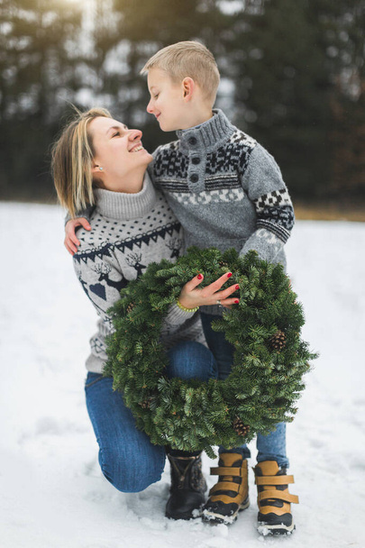 Cheerful mother and her son in gray knitted sweaters hold home made advent wreath, looking at each other, walking in winter forest or park outdoors - family together at christmas time - Photo, image