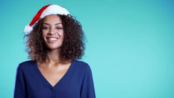 Copy space. Attractive mixed race girl in Santa hat celebrating Christmas or New year on blue background. Happy pretty african woman smiling and having fun. 4k - Video
