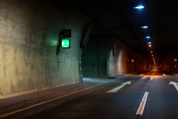 Automobiel auto donkere auto tunnel met witte pijlen op asfalt tonen weg richting. Nooduitgang bord met veel lichten. Lege ondergrondse weg. Stedelijke abstracte stad vervoer achtergrond - Foto, afbeelding