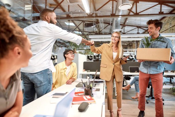 Nice to meet you. Young beautiful woman in suit giving fist bump to her new coworker while standing in the modern office with other colleagues - Foto, imagen