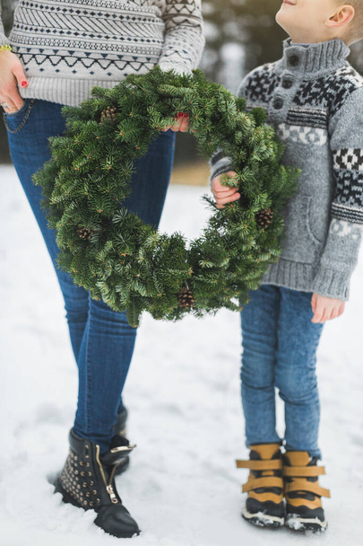 Cropped image of legs of mother and son, holding together beautiful Christmas wreath, standing outdoors in winter park or forest. Mother and child with Christmas wreath, outdoors - Photo, image