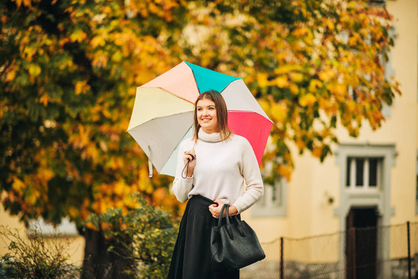 Yağmurun altında şık genç bir kadın, elinde renkli bir şemsiye, sıcak kazak ve siyah etek giyiyor. - Fotoğraf, Görsel