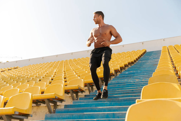 Image of healthy shirtless sportsman running by empty stadium se - Foto, afbeelding