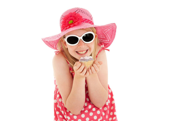 Happy, beautiful young blonde girl in big pink floppy hat and white framed sunglasses excitedly holding up a cup cake. Isolated on white studio background - Photo, Image