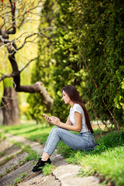 Attractive girl resting on nature at weekend. Girl in jeans clothes sitting near the lake in park. Caucasian brunette young woman sending message on smartphone outside alone.Technologies and gadgets.V - Foto, Bild
