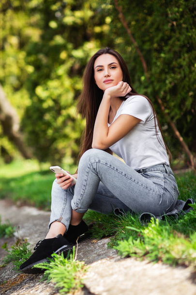 Attractive girl resting on nature at weekend. Girl in jeans clothes sitting near the lake in park. Caucasian brunette young woman with smartphone outside alone.Technologies and gadgets.Vertical - Photo, Image