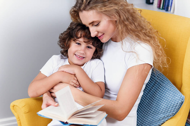 Bela mãe lê livro infantil para seu filho enquanto se senta em cadeira amarela confortável perto da janela na casa aconchegante
. - Foto, Imagem