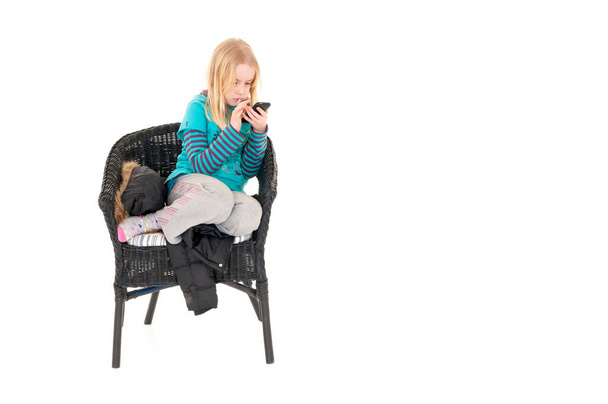 Młoda blondynka siedziała na wiklinowym krześle używając smartfona. Izolacja na białym tle studia - Zdjęcie, obraz