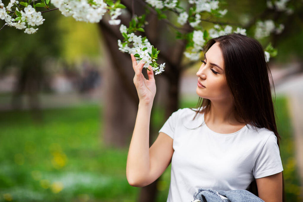 Girl standing under blossoming apple tree in natural park. Fashion concept. Elegant young lady in jeans clothes enjoying flowering garden at sunny spring day.Natural beauty makeup. Horizontal - Photo, image
