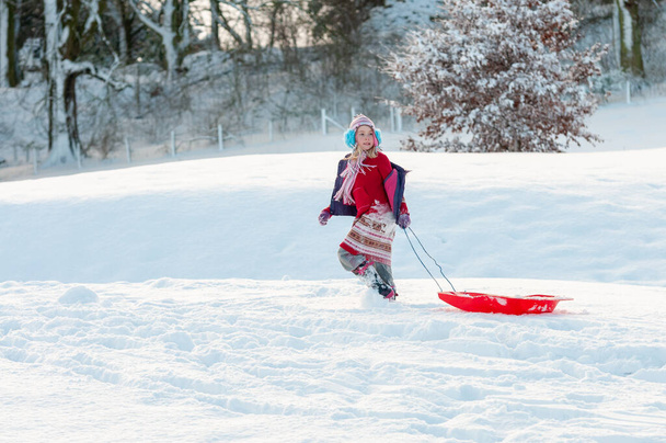 Chica joven vistiendo vibrantemente arrastrando su trineo detrás de ella a través de la nieve profunda en un entorno rural
 - Foto, imagen