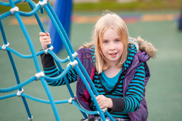Mooi jong blond meisje klimmen op een touw ladder in een speeltuin - Foto, afbeelding