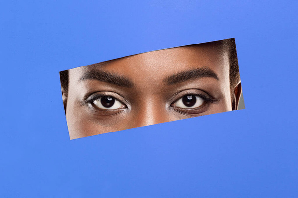 Afro naiset silmät ilman meikkiä kurkistaa reiästä sininen paperi
 - Valokuva, kuva
