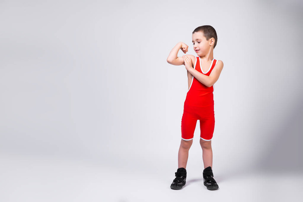  The concept of a little fighter athlete. Little funny boy in a red sports ortsovskoy leotard and wrestling boots watches his muscles grow on his arms and smiles at his achievements on a white isolated background - Φωτογραφία, εικόνα