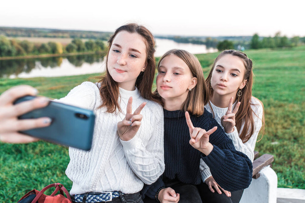Three teenage girls, teenagers in summer city have rest after school college, hands smartphone, selfie picture video call online application on social networks. Happy smiling. Casual clothes, sweater. - Фото, изображение