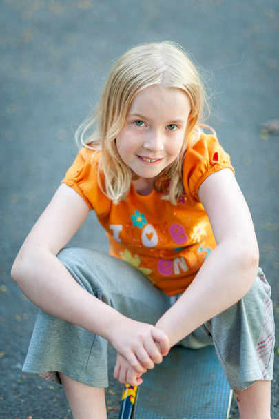 Счастливая, красивая блондинка, улыбающаяся и сидящая на скейтборде при зрительном контакте
 - Фото, изображение