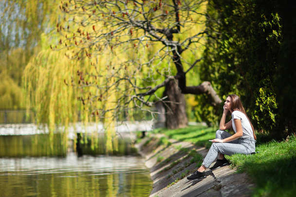 Attractive girl resting on riverbank near forest at spring warm day. Relaxing on nature at weekend. Girl in jeans clothes sitting near the lake in park. Nature concept. Fashion photoshoot. Horizontal. - Foto, Imagen