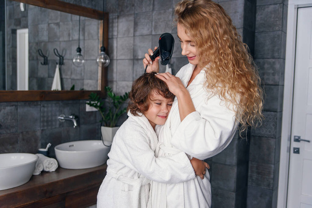 Mom and son have fun together in the bathroom. Beautiful mother with her little son dressed in bathrobe are relaxing and playing in the bathroom together. - 写真・画像