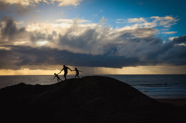 Silhouette of man and children on beach at sunrise with storm visible in the distance - Foto, afbeelding
