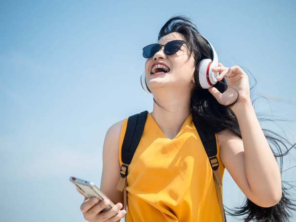 Mujer asiática feliz con mochila usando auriculares en la playa, li
 - Foto, imagen