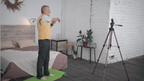 atractivo anciano muestra ejercicios de bienestar grabando videon smartphone para redes sociales y seguidores en acogedora habitación con interior de diseño
 - Metraje, vídeo