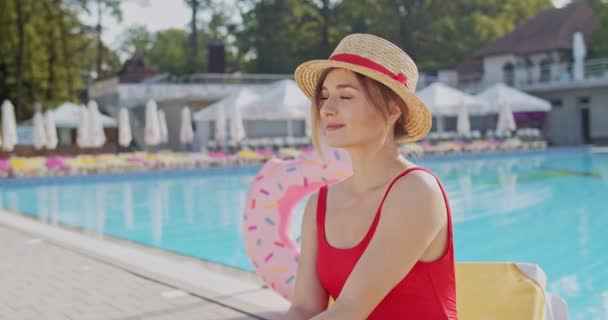 Portrait shot of the young Caucasian pretty and happy woman in hat and red swimming suit relaxing at the swimming pool outdoors and smiling to the camera. - Кадры, видео