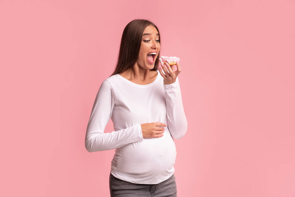 Беременная леди кусает пончик на розовом фоне, студия
 - Фото, изображение