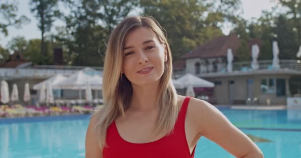 Portrait of the yung pretty blonde Caucasian woman in the red swimming suit smiling to the camera at the swimming pool at the resort. Close up. - Кадры, видео