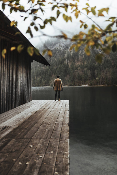 Πλήρης φωτογραφία σώματος νεαρού άνδρα φωτογράφου με καφέ παλτό που τραβά φωτογραφίες με μια μικρή κάμερα χωρίς καθρέφτη να στέκεται σε ένα κατάστρωμα σπιτιού στη λίμνη Plansee, Αυστρία - Φωτογραφία, εικόνα
