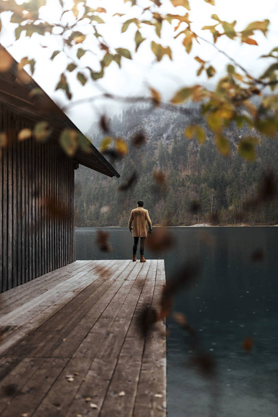 Full body shot of young male photographer in brown coat taking pictures with a small mirrorless camera standing on a lake house deck in lake plansee, Austria - Фото, изображение