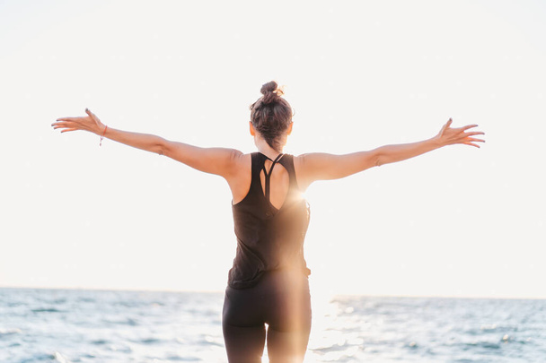 Slim woman in black bodysuit practicing yoga near sea or ocean during sunrise light. Flexibility, stretching, fitness, healthy lifestyle.  - Zdjęcie, obraz