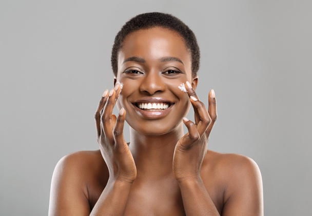 Portrait de heureuse femme noire appliquant crème hydratante sur le visage
 - Photo, image