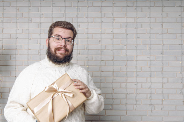 Boże Narodzenie koncepcja środkowy dorosły mężczyzna portret w okularach z prezentem w rękach patrząc na aparat fotograficzny na białej ścianie cegły proste tło puste miejsce na tekst tutaj wzór reklamowy  - Zdjęcie, obraz